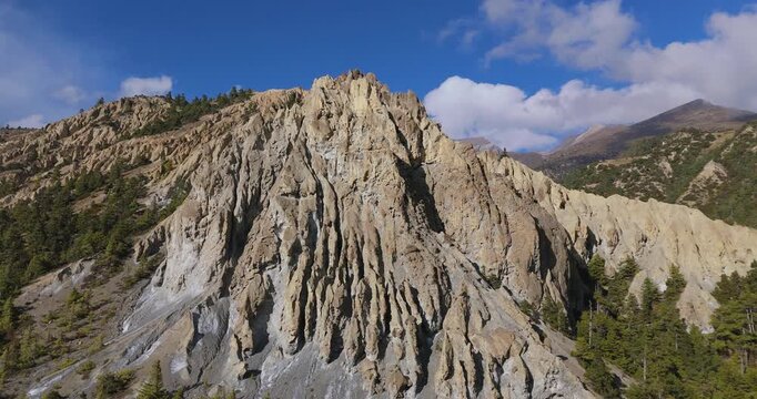 Annapurna Circuit, Manang region, Nepal, aerial cruise shot of a massive eroded rocky cliff face surrounded by green pine forests beneath a blue sky with white clouds.