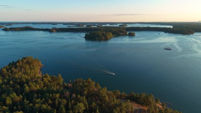 Aerial view of a motorboat leaving a long V-shaped wake across calm blue waters of the Helsinki archipelago at golden hour, with forested islands and distant city skyline