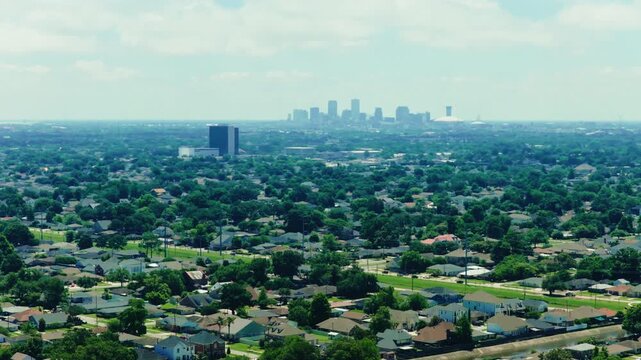 Aerial over Metairie in Jefferson Parish, Louisiana, looking across tree-covered residential blocks toward the New Orleans downtown skyline rising through summer haze.