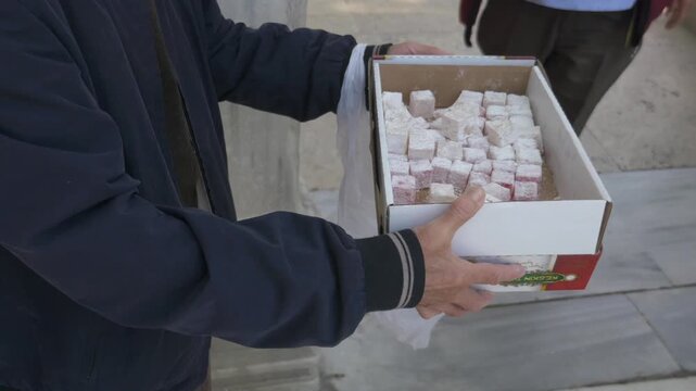 Close up of person holding box of turkish delight sweets while people take pieces in courtyard outdoors