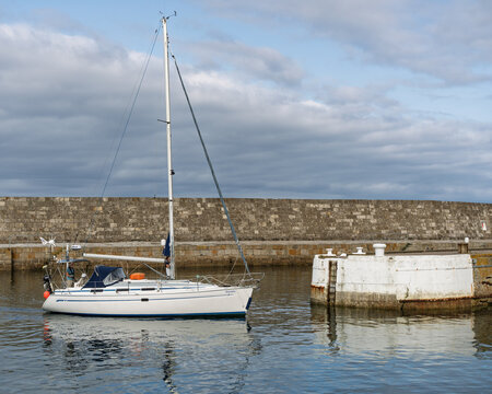 Sailing boat entering Lossiemouth Harbour in Moray Scotland during the calm afternoon with a stone wall backdrop and cloudy sky overhead