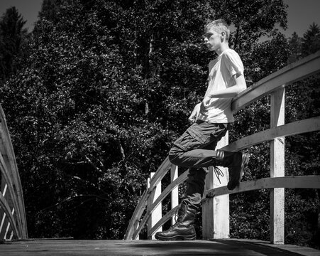 Black and white photo of a teenage boy standing on a curved wooden bridge, leaning on the railing in casual clothing with trees in the background