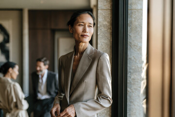 Fototapeta premium A poised woman stands by a large window, exuding confidence in a stylish suit. Her thoughtful gaze reflects determination and grace in a modern corporate environment.