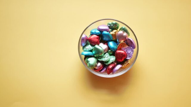Top view of hand picking heart shaped chocolate from glass bowl on yellow background