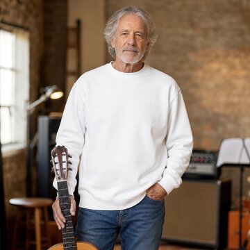 Musician stands in a music studio holding an acoustic guitar with amplifiers in the background.