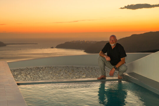 Man kneeling beside an infinity pool overlooking Santorini&rsquo;s cliffs and sea at sunset, capturing serene Mediterranean evening light.