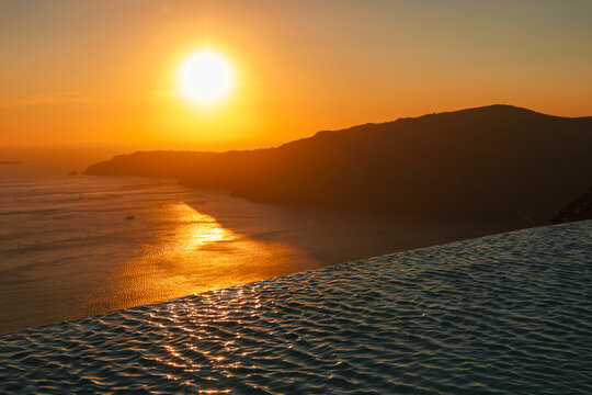 Golden sunset reflecting on calm infinity pool water overlooking Santorini&rsquo;s cliffs and the tranquil Aegean Sea.