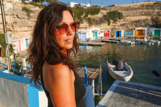   
Smiling woman in sunglasses standing by the colorful seaside boathouses of Mandrakia, Milos, Greece, captured in bright summer light.