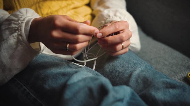 Close-up of an anonymous young woman's hands sitting on a sofa at home, trying to untangle wired earphones before giving up and switching to wireless earbuds for a more convenient listening experience