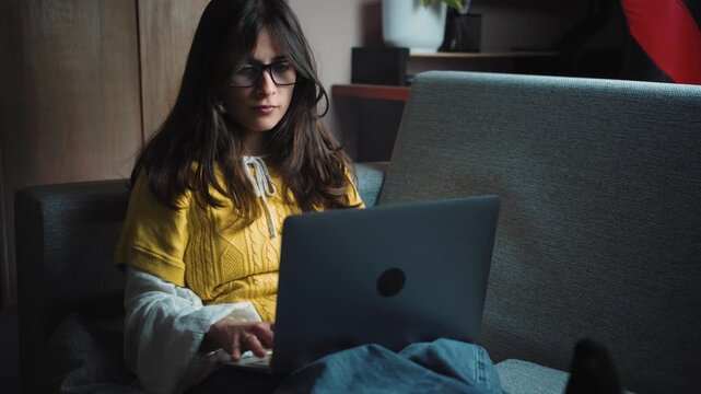 Young Hispanic woman working on her laptop while relaxing on the sofa at home. Cozy remote work lifestyle scene representing productivity, digital work, freelancing and work-life balance.
