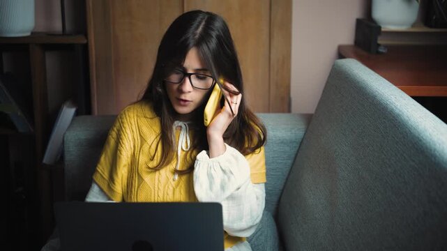 Young Hispanic woman talking on the phone while entering data on her laptop from the sofa at home. Remote work lifestyle scene representing multitasking, productivity, technology and digital business.