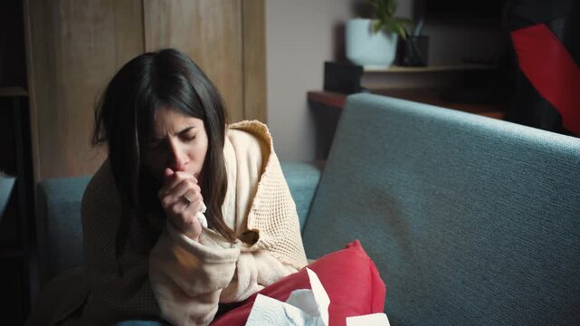 Young Hispanic woman sitting on her sofa at home with severe cough and fever. Health lifestyle video representing flu symptoms, illness, recovery, self-care and the need for rest.
