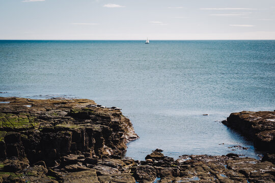Whitley Bay UK: 13th April 2025: A sailboat glides over the water near the coastline of Whitley Bay on a clear, sunny day. The rocky shoreline frames the scene as the sea stretches toward the horizon