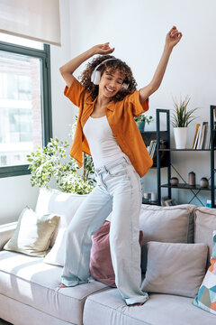 Motivated happy woman singing and dancing on couch while listening music with headphones at home