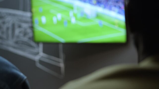 People gathered in the living room watching a soccer match on TV. View from behind of a group of young adults enjoying a football match on television, sharing snacks and drinks in a cozy living room.