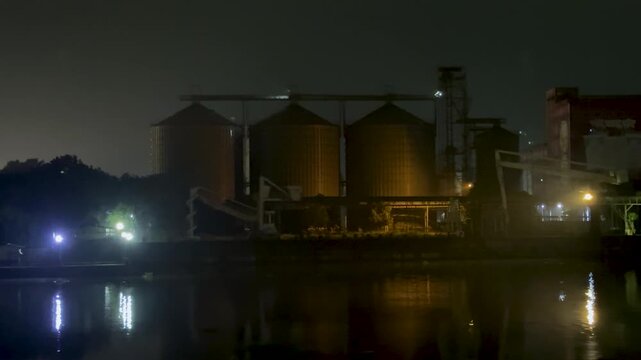 Industrial silos at night with reflections on a calm river under a dimly lit, cloudy sky