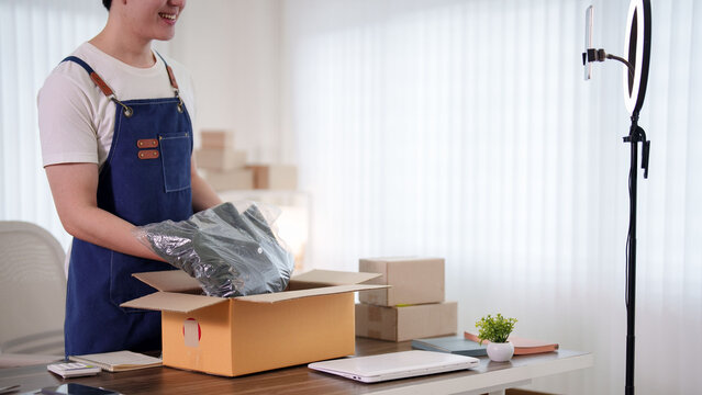 Young Asian online seller packing wrapped product into shipping box at home workspace with ring light