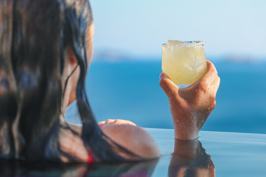 A woman enjoys a chilled cocktail while relaxing in an infinity pool overlooking the blue sea and distant islands under bright Mediterranean sunlight on Santorini in Greece.