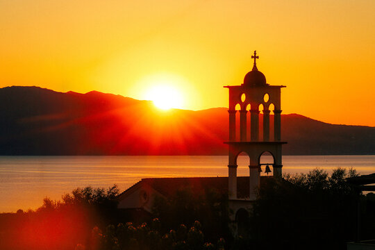 A church bell tower in Kaliviani village near Kissamos, Crete, silhouetted against a glowing Mediterranean sunrise over the sea, evoking peace and timeless island beauty.