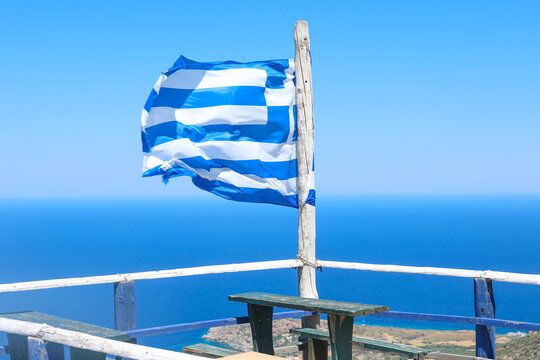 A Greek flag waves proudly in the wind from a rustic terrace overlooking the deep blue sea in Crete, capturing Mediterranean color and coastal serenity.