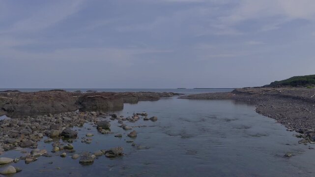 Flying over glassy blue water as coastal river meets calm ocean on rocky beach