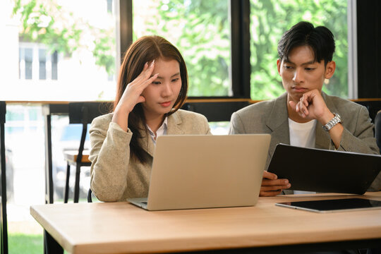 Two Young Asian Business Professionals Looking Stressed While Reviewing Work on Laptop and Tablet in Modern Office