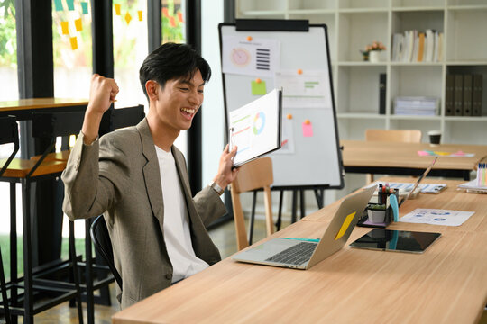 Excited Young Asian Businessman Celebrating Success While Reviewing Business Charts and Reports in Modern Office