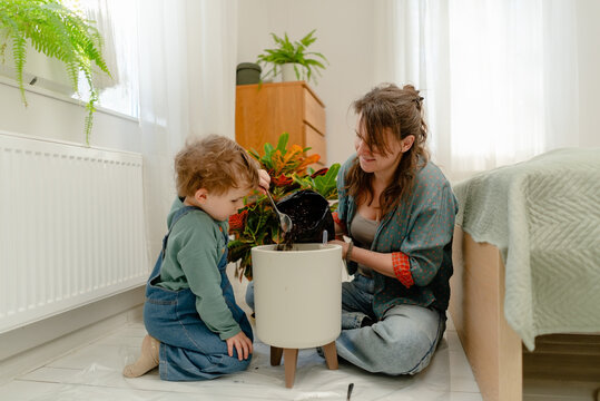 A calm mother holds a potted plant and reaches for leaves on a bright sunny floor. Her curious son watches while the warm mom smiles caring for indoor plant greenery in a cozy home.