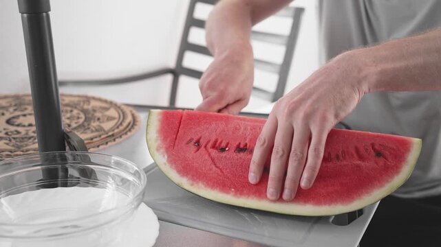A person cuts a watermelon on a kitchen table. The scene shows the steps of holding the fruit and slicing it with a knife. It is bright and focuses on the cutting process.