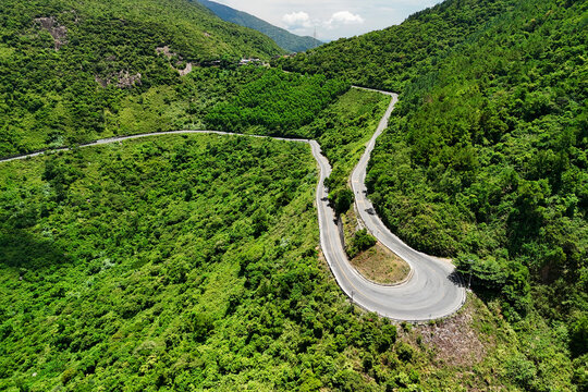 Asphalt road winds through lush green mountains on Hai Van Pass in Vietnam during a clear sunny afternoon while vehicles travel across the steep tropical landscape