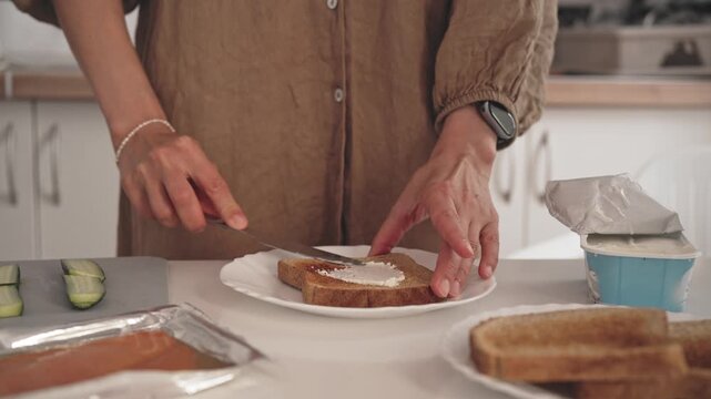 A person prepares breakfast by spreading butter on a slice of toast in a kitchen. The action shows hands using a knife to apply the butter evenly on the bread.