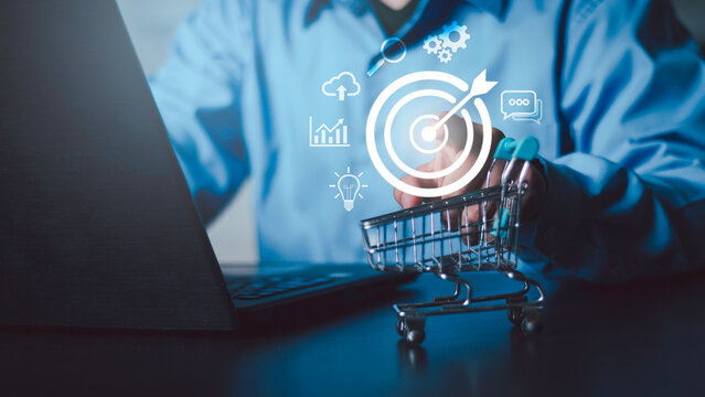 Businessman pointing at glowing target board with shopping cart and marketing icons. E-commerce strategy, customer targeting, business goals, and digital data analysis on laptop background.