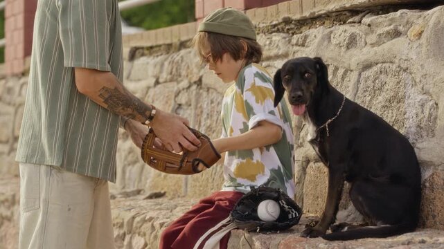 Caucasian father putting baseball glove on young sons hand while sitting outdoors next to black dog. Clip is ideal for family bonding, parenting, and youth sports advertisements