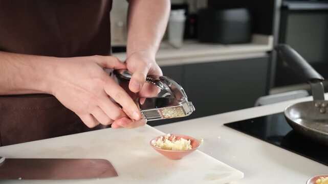 Man's hands using a garlic press to crush a clove into a small bowl on a white cutting board