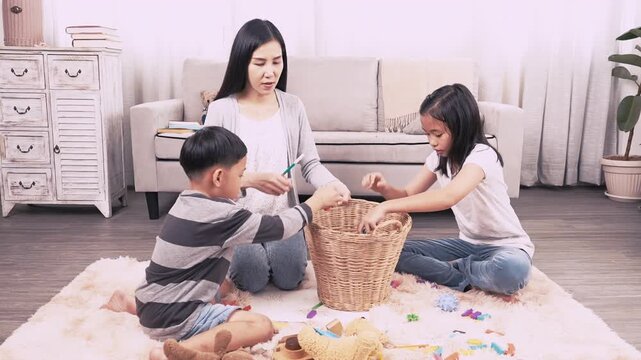 Asian mother sits on rug in living room with son and daughter, happily keeping together with colorful toys. They share a sweet family moment as they play near a wicker basket, loving childhood memory