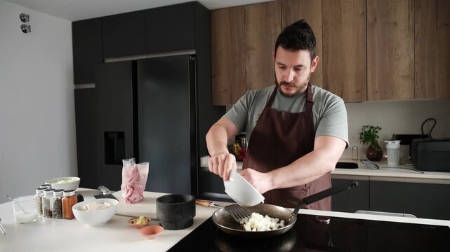 Young man wearing an apron preparing food by frying chopped onions in a pan on an induction hob