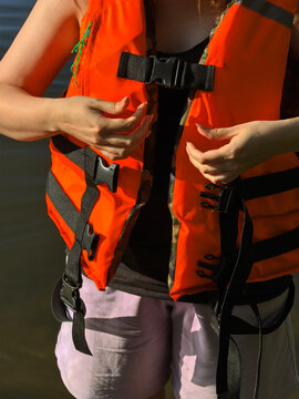 Woman putting on unbuckled bright orange life vest standing near water front view, demonstrating personal flotation device fitting step, safety gear tutorial, and water activity preparation.