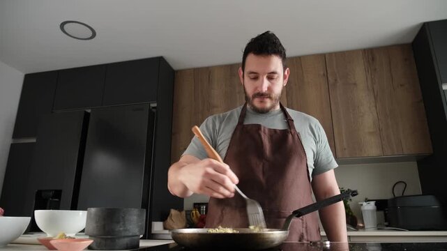 Millennial man wearing an apron stirs steaming risotto in a frying pan on an induction hob