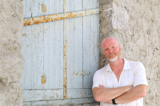 Natural light portrait of a man with a beard and white shirt standing confidently against a textured stone wall and weathered blue shutter, evoking rustic charm and authenticity.
