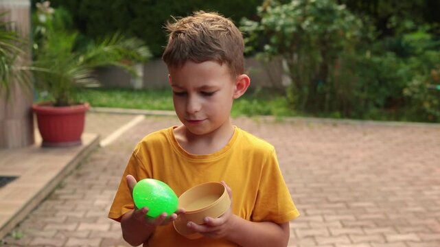 A boy holds a sparkly green dumpling toy in his hands, squeezing its squishy glittery surface while standing outdoors in natural daylight