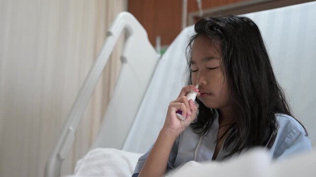 Young Asian child patient resting in a hospital bed using a nasal spray to treat sinus allergy symptoms and breathing problems during a childhood sickness recovery inside a private clinic.