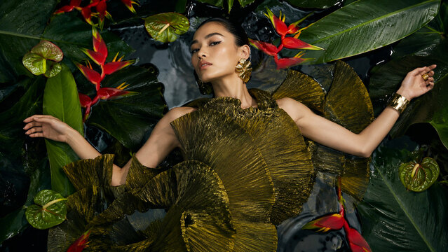 Editorial fashion portrait of a woman in an olive couture dress surrounded by tropical leaves and red exotic flowers. Ideal for luxury, beauty, botanical and advertising use.