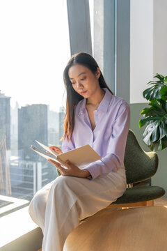 Asian Woman Reading a Book by the Window in a City Office