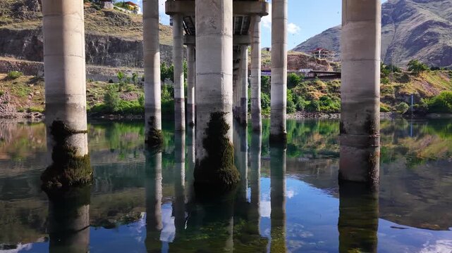 Smooth cinematic flight moves from right to left beneath towering concrete bridge piers rising from calm water, showcasing weathered textures, moss-covered surfaces, and impressive structural scale.