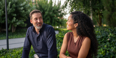 Fototapeta premium A cheerful couple sits on a park bench, relax and each others company on a sunny day. The scene captures relaxation and happiness in a lush green environment.