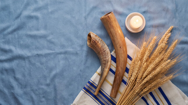 A traditional Jewish holiday scene featuring two shofar horns, a tallit prayer shawl, golden wheat stalks, and a lit candle on a soft blue textured background.