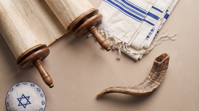 A high-angle view of traditional Jewish religious items including a Torah scroll, a shofar, a tallit prayer shawl, and a kippah on a neutral background.