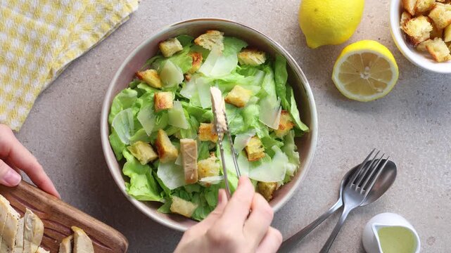 Top view of a hand adding grilled chicken pieces to a fresh Caesar salad with iceberg lettuce, croutons and Parmesan cheese. Flat lay food preparation on a beige table. Horizontal video.