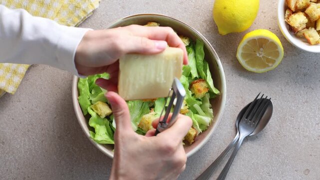 Top view  of a hand shaving fresh Parmesan cheese over a homemade Caesar salad with crisp iceberg lettuce and croutons. Flat lay food preparation on a beige table. Horizontal video.