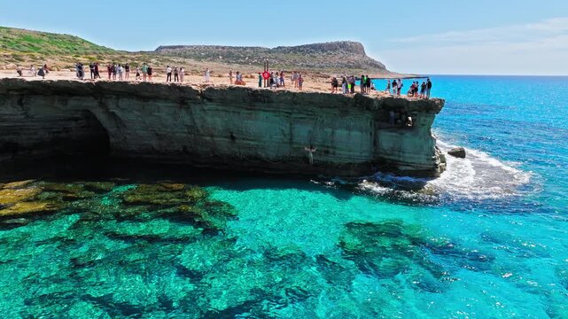 Aerial view of Cape Cavo Greco sea caves in Cyprus. Tourists visit the rocky cliff edge above crystal-clear turquoise water, natural cave openings and rugged Mediterranean coastline.